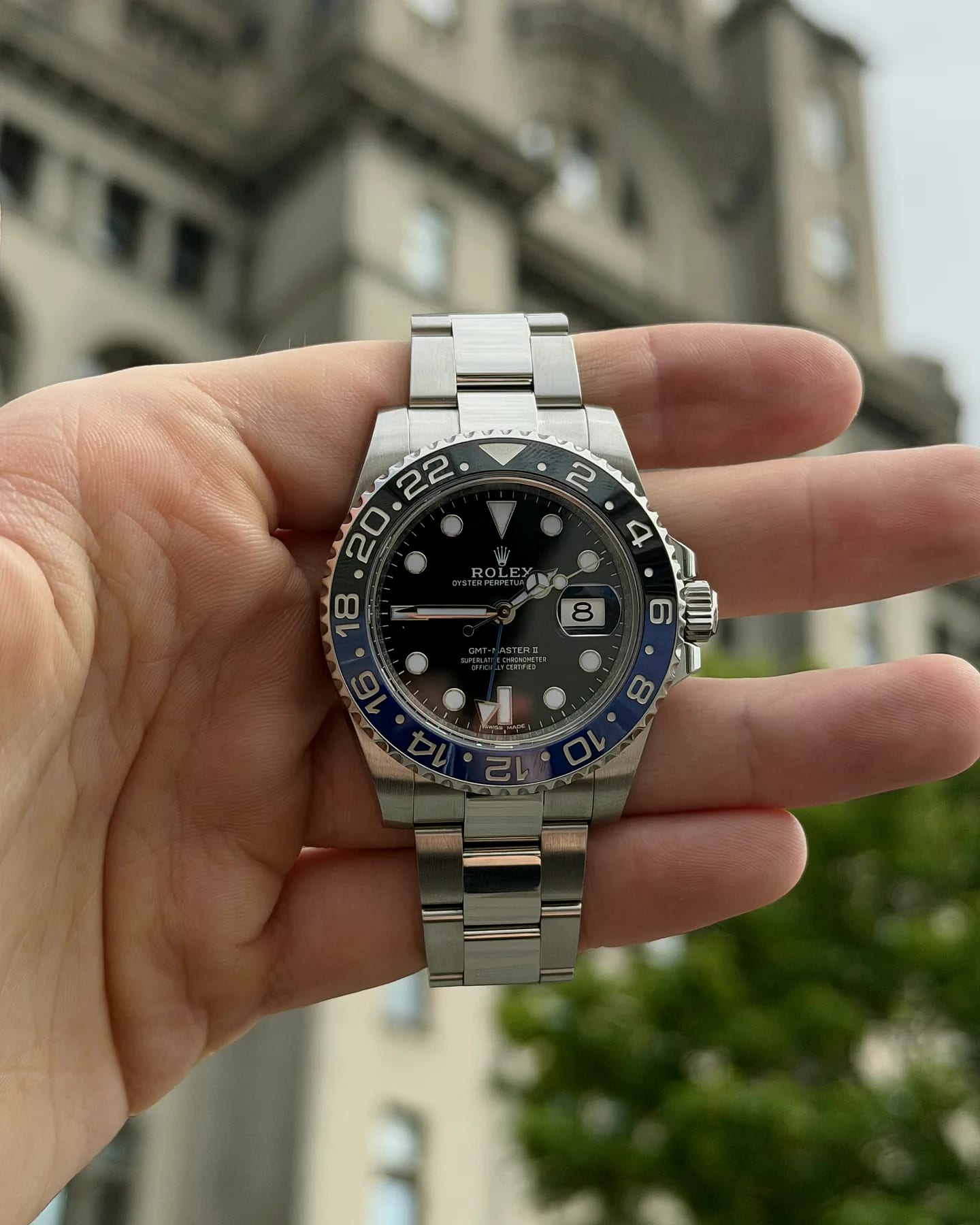 A Rolex GMT-Master II with a black and blue Batman bezel, held in an open palm outdoors with a period stone building behind